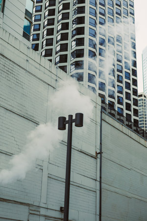 Steam is coming out of a vent next to a tall building in a city. The sky is clear and the setting is...