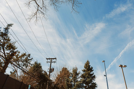 The sky shows clouds with trails and blue patches. Trees are present below with power lines running...