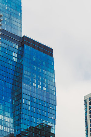 People walk around as tall buildings with blue glass reflect the sky on a cloudy day in a busy urban...