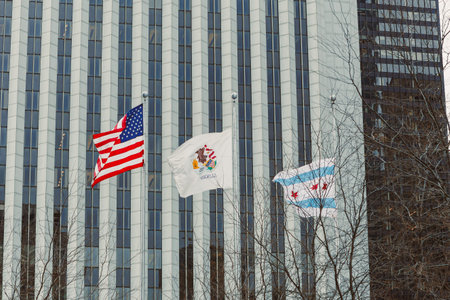 Three flags wave in front of a tall building while bare trees stand nearby on an overcast day in a...