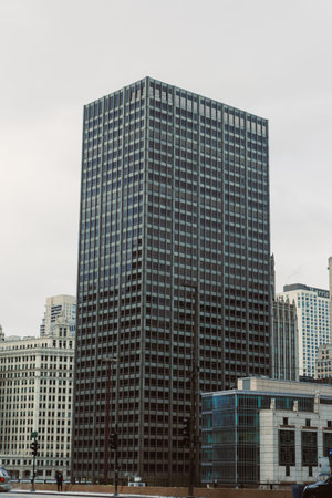A tall office building rises above nearby structures in a busy city. The sky is gray and clouds are...