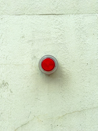 A red circular light is mounted on a plain white wall. The light serves as a warning or alert. It is...