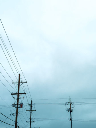 Power lines connect tall poles as they stretch across the cloudy sky in an urban area during the...