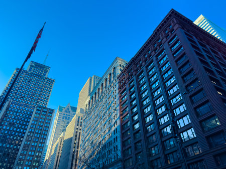 Tall buildings stand in an urban setting under a clear blue sky. A flag flutters in the foreground...