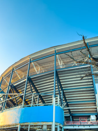 Workers build a new stadium with a large metal frame and blue sections. The sky is clear, and the...