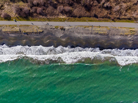 Waves hit the shore with foam while a road runs alongside the beach. People walk on the path close...