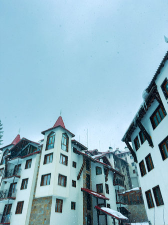 Snow is falling on a mountain resort with several white buildings. The scene shows a gray sky and a...