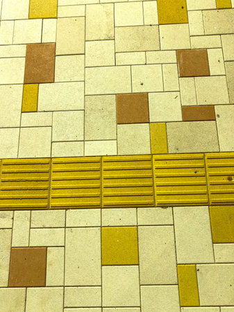 Tiles in various colors cover a sidewalk with a yellow textured strip guiding visually impaired...