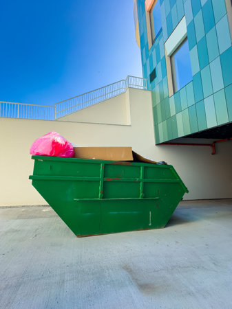 A green dumpster stands in an urban area. It is filled with pink bags and cardboard boxes. A...