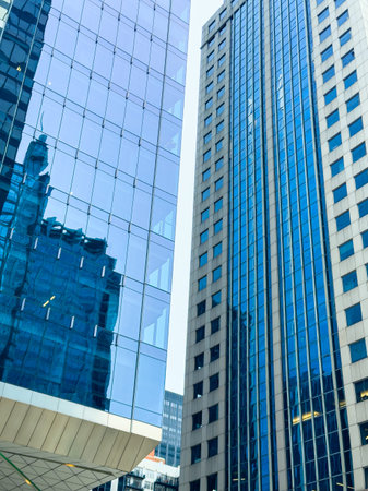 Tall buildings with glass walls reflect the blue sky in a busy city. Sunlight shines on the...