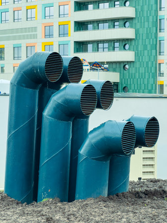 Five ventilation pipes rise from the ground at a construction site near a new building. The area is busy with machinery and other construction materials.の写真素材