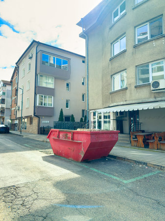 A red dumpster sits on a street in front of buildings. There is a clear sky and visible commercial...