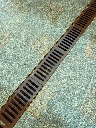 Water flows through a metal drainage grate set in a stone pavement. The scene is typical of a city...