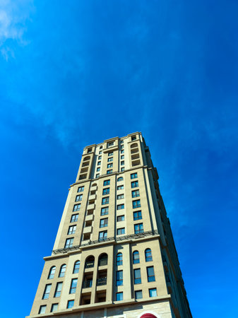 A tall building stands in an urban area. The structure has many windows. The sky above is blue with...