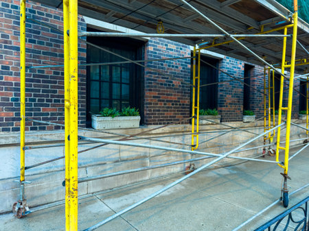 Workers are setting up scaffolding around a building. The brick wall shows window planters at the...
