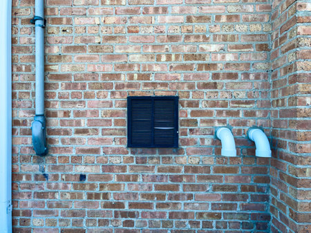 A brick wall displays a vent and two white pipes located near the side of a building in bright...
