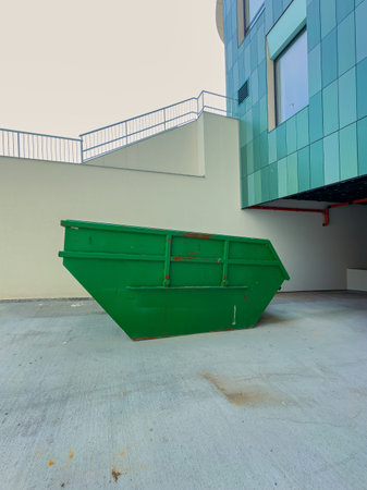 A green dumpster stands in a concrete space next to a modern building. The area is empty, and there...