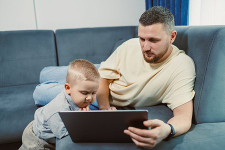 A father sits on a couch with a laptop while his young son leans over to look at the screen. They...