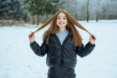 A young woman stands in a snowy park holding her long hair while smiling. She wears a warm jacket...