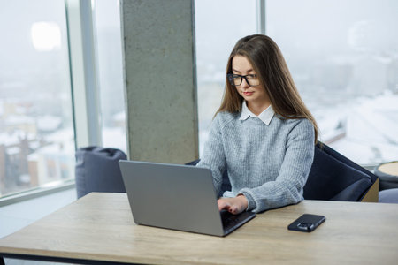 A woman sits at a wooden table in an office. She writes on her laptop while wearing a sweater and...