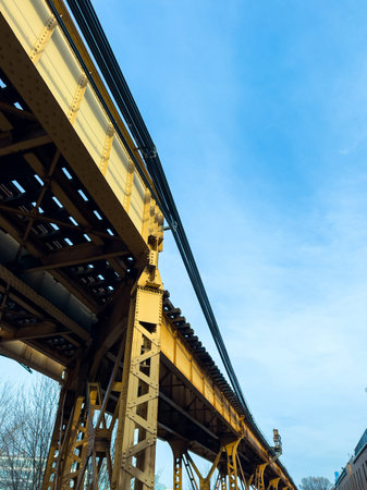 Elevated train tracks stretch across the scene showing steel beams and wires. Blue sky and trees are...