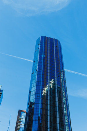 A tall skyscraper stands under a clear blue sky. The glass facade reflects other buildings nearby. A...