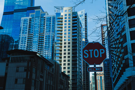 A stop sign stands at a city intersection surrounded by tall buildings. Sunlight reflects off the...