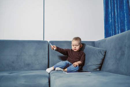 A young child sits comfortably on a blue couch in a living room. The child is focused on the tablet...
