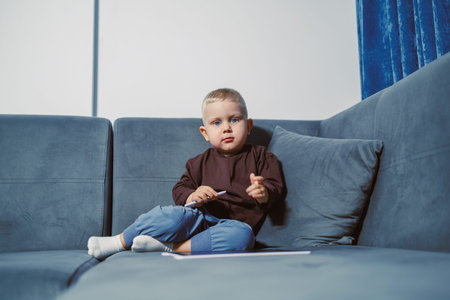 A child sits on a blue sofa, holding a pen and looking directly at the camera. Papers are on the...