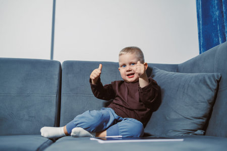 A young child sits on a blue couch, shows thumbs up with both hands, and smiles. The room has a...
