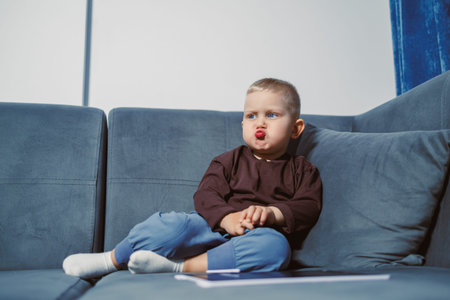 A young child sits on a blue couch making a silly expression. The room is bright, and the child...