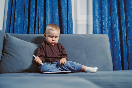 A young child sits on a couch in a living room and looks at a tablet while holding a stylus. The...