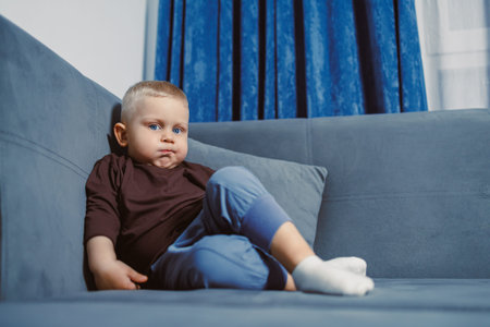 A young child sits on a blue sofa with a serious face. The living room has blue curtains and the...