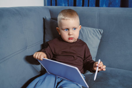 A young child sits on a blue couch in a living room while holding a tablet and a stylus. The child...
