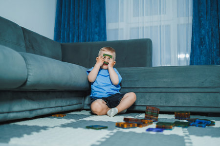 A young child sits on a rug on the floor in a living room. The child holds a building block in front...
