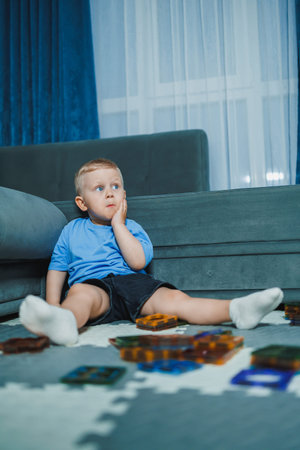 A young boy is sitting on a carpet in a living room. He has a thoughtful expression as he looks at...