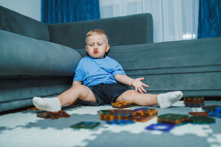 A young boy sits on the floor of the living room surrounded by colorful building blocks. He looks...