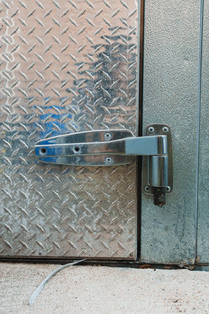 A metal handle and lock are fixed to a door made of diamond plate. The surroundings show a concrete floor and the texture of the door.