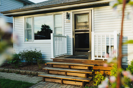 A house has a wooden porch with stairs leading to the front door. The porch is white and there are flowers growing near the entrance. The sun is low in the sky.
