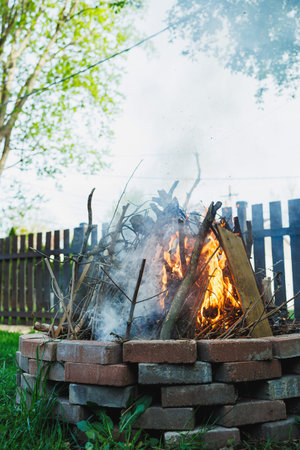 A campfire burns brightly in the backyard. Smoke rises from the fire as sticks and wood crackle. The sunlight shines on the green grass and wooden fence nearby.
