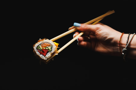 A person holds a sushi roll with chopsticks in one hand. The background is dark. The sushi displays colorful ingredients like fish and vegetables. This happens during a meal.