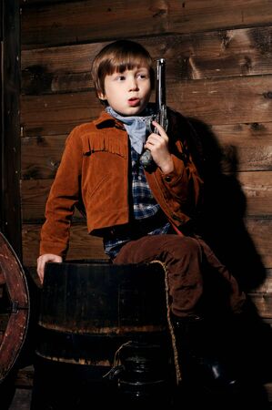 studio portrait of little boy wearing cowboy costume and sitting on a wooden barrel in wooden...