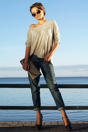 beautiful girl standing against blue sea and sky and looking at you. Young stylish woman in pullover and jeans with handbag. Summer outdoor fashion portrait.の写真素材