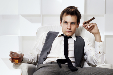 Young rich successful man sitting in white leather armchair with cigar and glass of whiskey. Fashion style portrait in modern interior.の写真素材