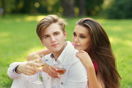 Young beautiful caucasian couple hugging and drinking wine at picnic in summer park on green grassの写真素材