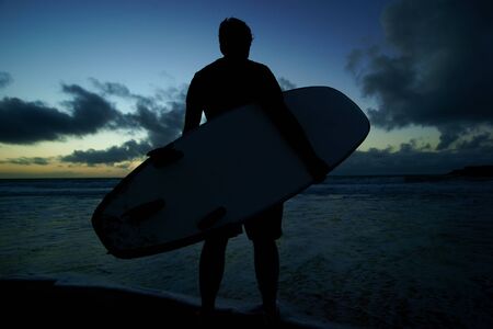 Silhouette of young male surfer standing at the beach and holding surfboard at sunset against blue sky with clouds backgroundの写真素材