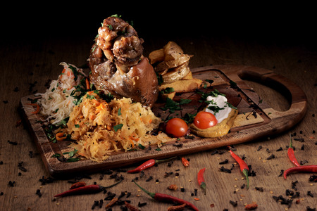 Grilled pork knuckle served with braised cabbage, fresh cherry tometoes and mustard served on wooden board on rustic wooden counterの写真素材