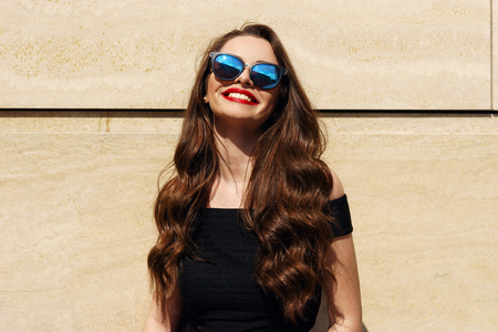 Closeup portrait of young happy smiling beautiful girl with long brunette curly hair posing against wall and smiling on a sunny warm dayの写真素材