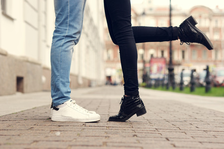 Close up of legs of couple kissing at city street with stone pavement. Man in blue jeans and white sporty sneakers and woman in black trousers and black leather bootsの写真素材