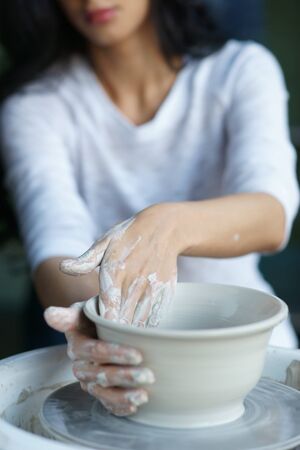 Young beautiful pretty woman with brunette dark hair working on pottery wheel and sculpting clay pot. Shallow DOF. Focus on handsの写真素材